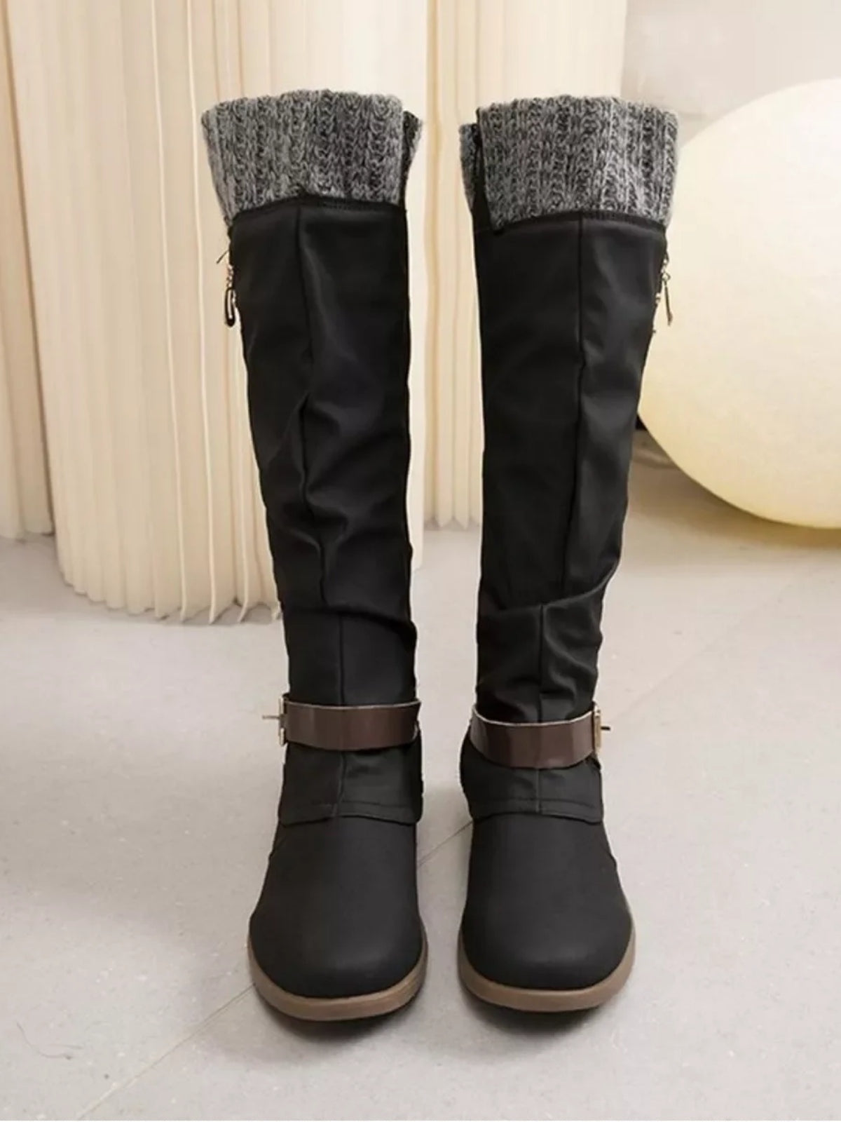 Black knee-high slouch boots feature gray knit cuffs, low heels, ankle straps, relaxed shafts, and contrasting rubber soles.