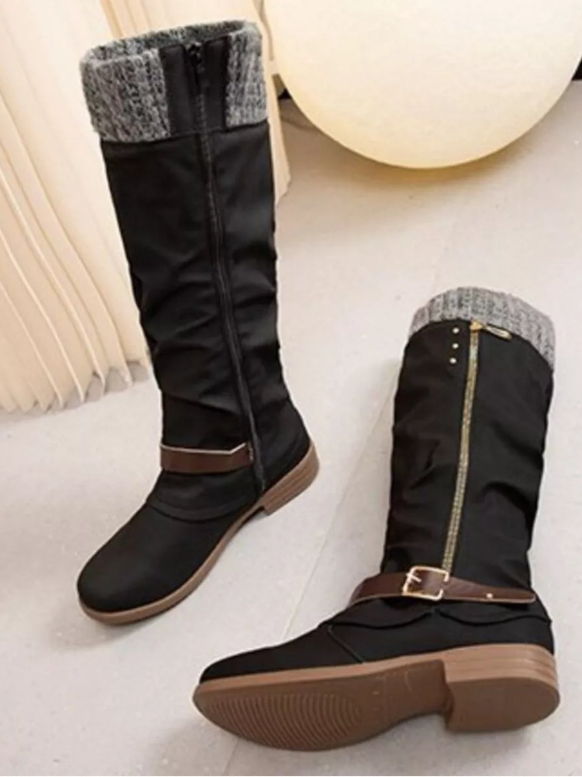 Black knee-high slouch boots feature gray knit cuffs, low heels, brown buckle straps, side zippers, and durable rubber soles.