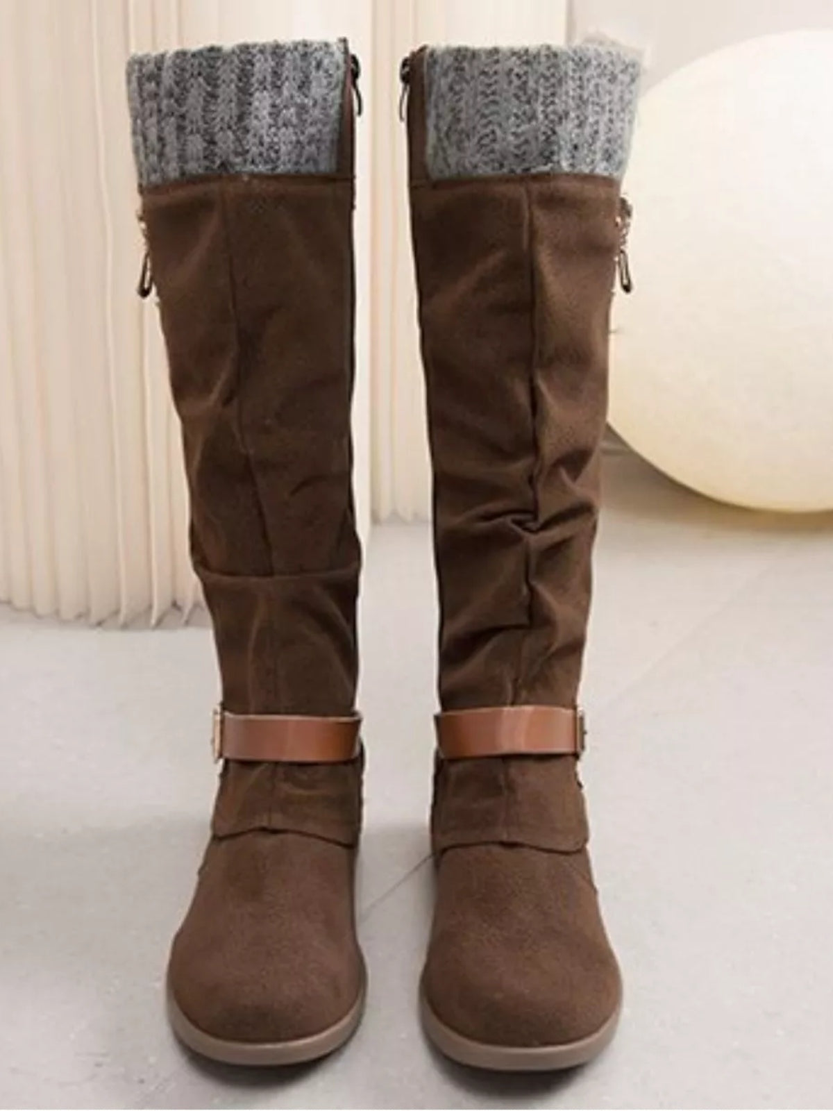 Brown suede knee-high slouch boots feature gray knit cuffs, low heels, rounded toes, and strap details around the ankles.