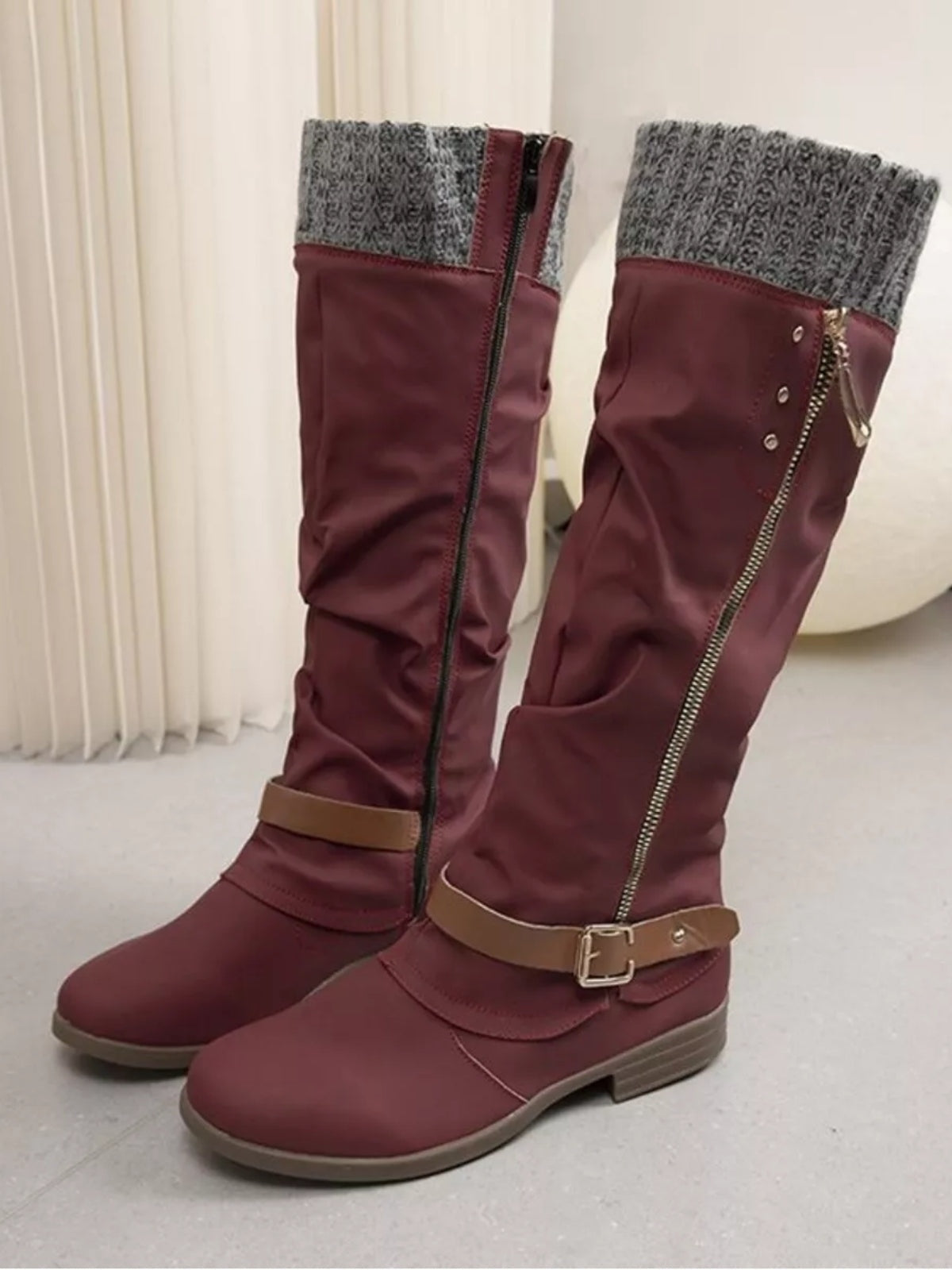 Burgundy knee-high slouch boots feature gray knit cuffs, low heels, side zippers, and buckled ankle straps over a rounded toe.