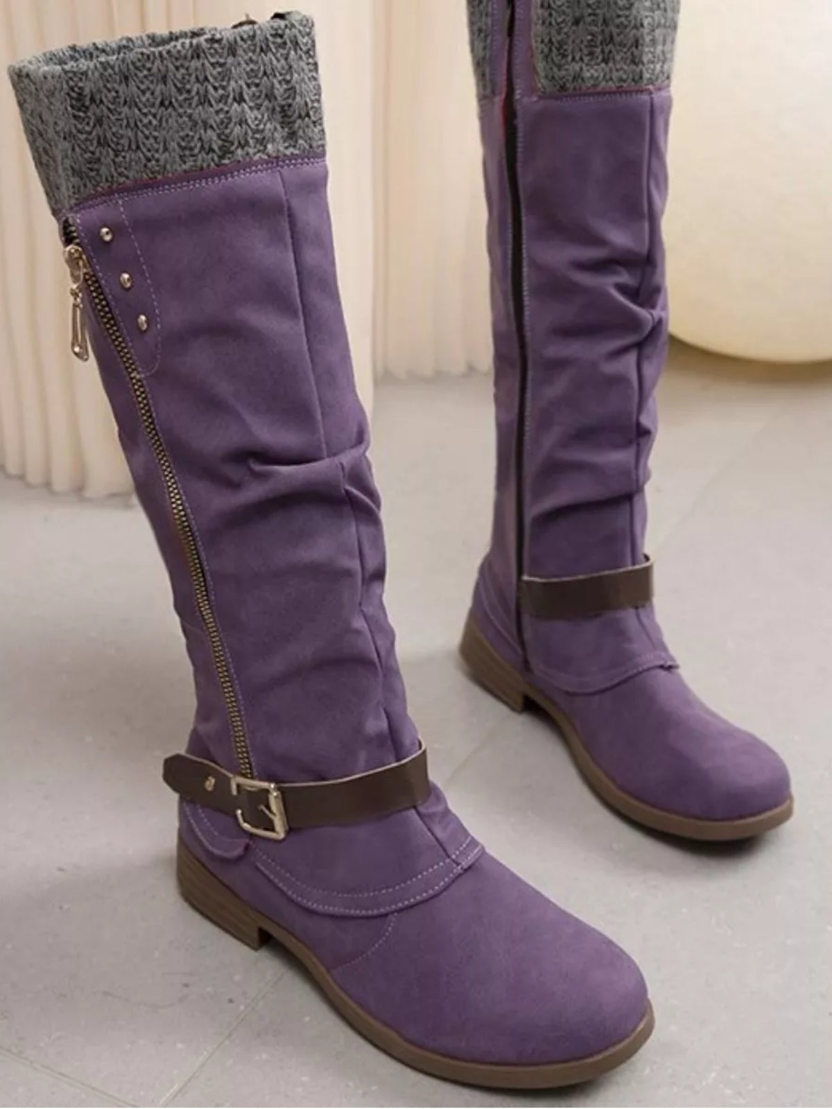 Purple suede knee-high slouch boots feature gray knit cuffs, low heels, side zippers, ankle buckles, stitched panels, and rubber soles.