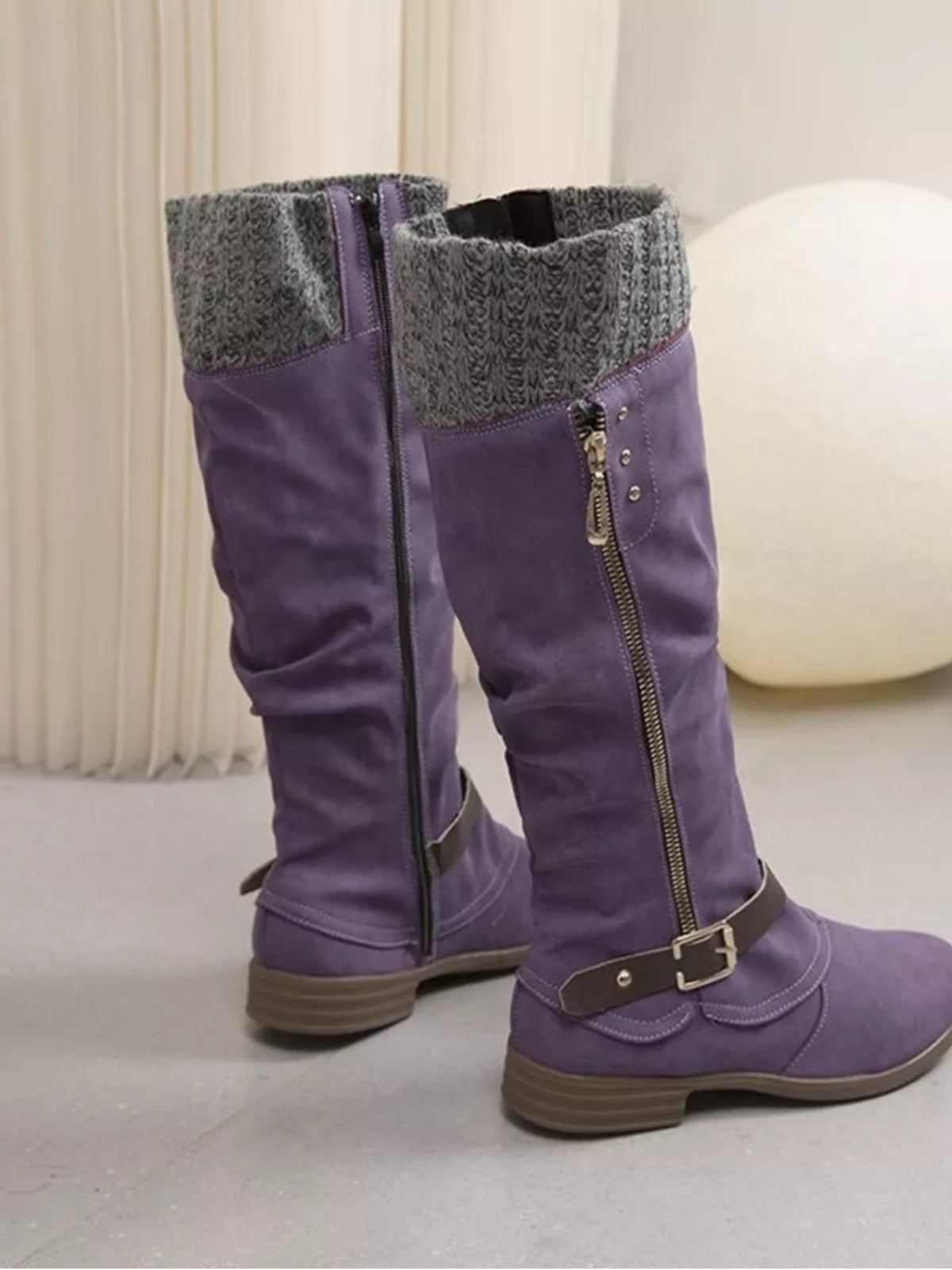 Purple suede knee-high slouch boots feature gray knit cuffs, low heels, side zippers, decorative buckled straps, and sturdy rubber soles.