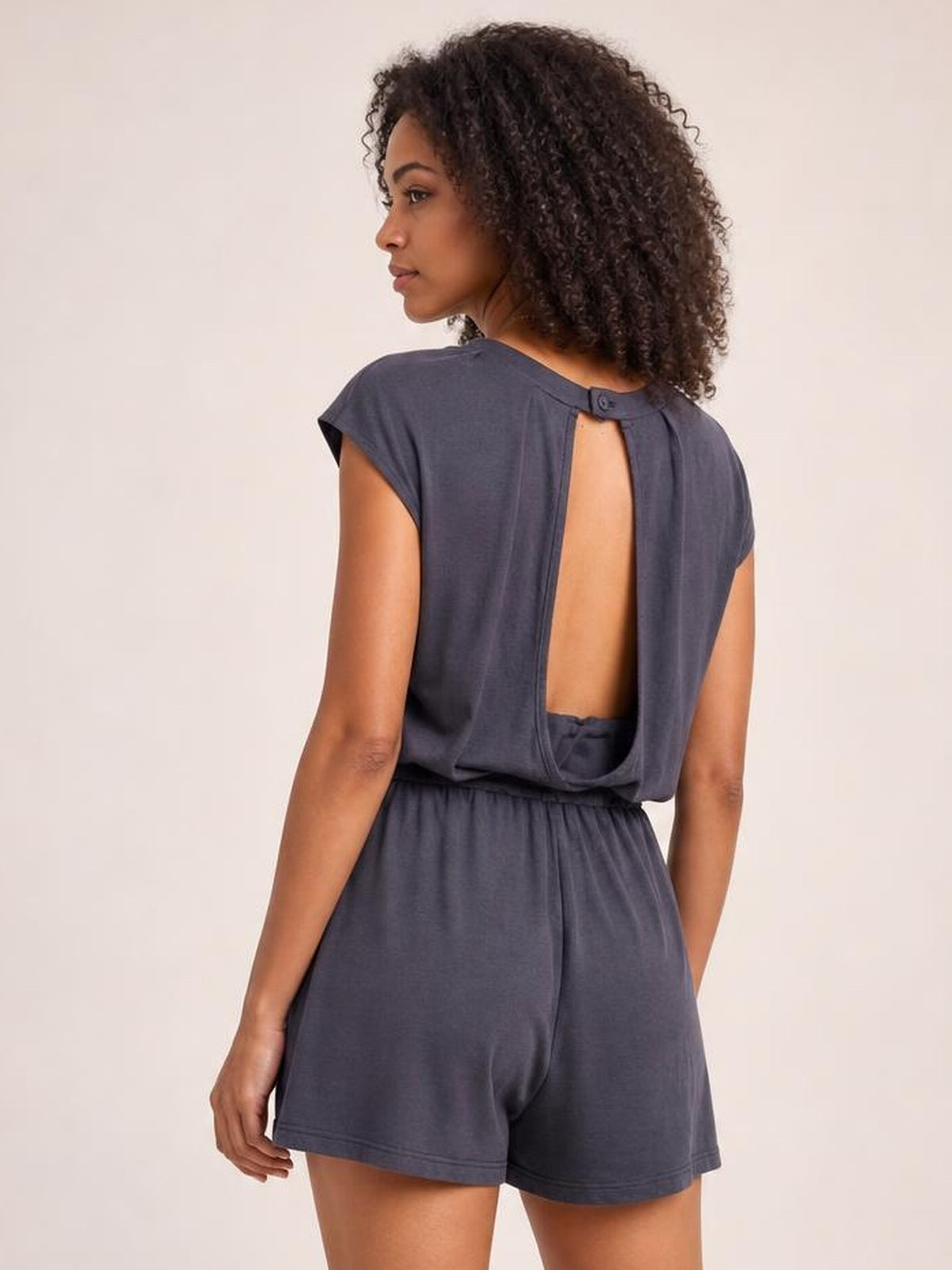 Model shown from the back in a charcoal knit romper with cap sleeves, an open-back cutout, adjustable drawstring waist, and relaxed shorts silhouette.