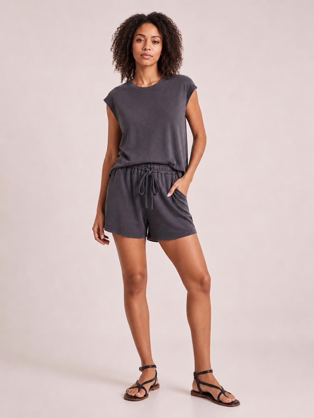 Model wearing a charcoal knit romper with cap sleeves, a relaxed silhouette, elastic drawstring waist, and tailored shorts, standing with one hand in a pocket.