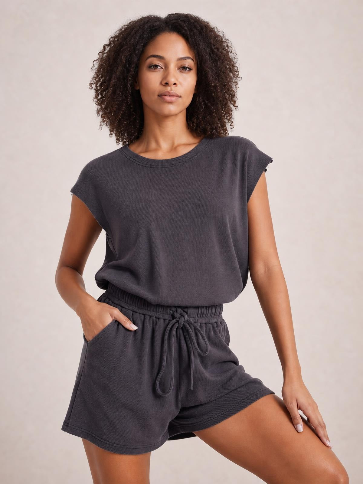 Model wears a charcoal knit romper with cap sleeves, a relaxed silhouette, elastic drawstring waist, and loose shorts, posing with one hand in a pocket.