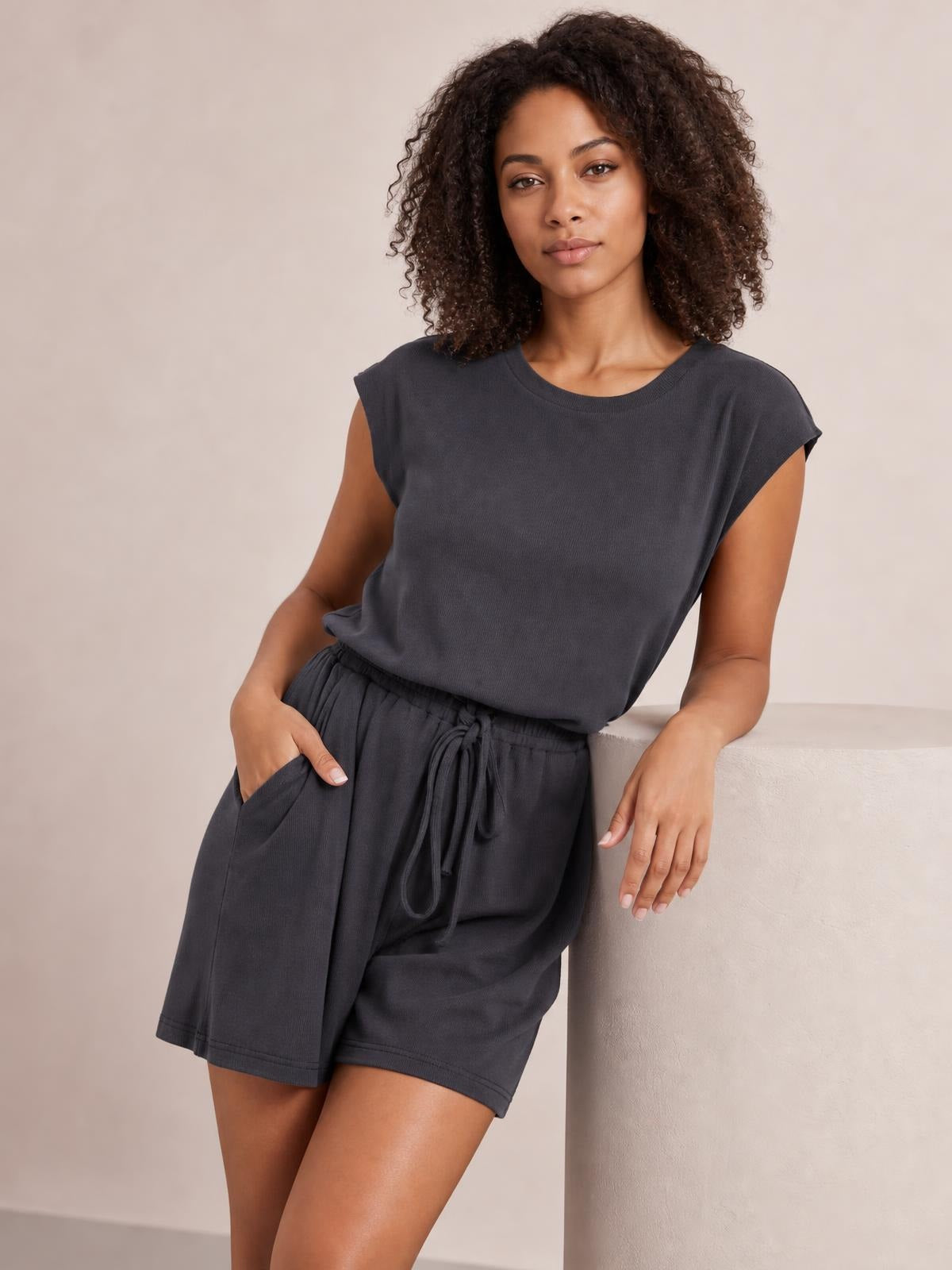 Model wears a dark charcoal knit romper with cap sleeves, a crew neckline, relaxed shorts, and an adjustable drawstring waist, leaning with one hand in a pocket.