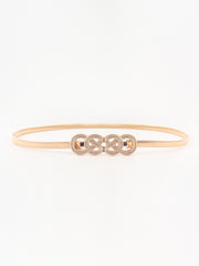 Slim gold-tone metal belt with a subtly textured band and ornate interlocking knot buckle, shown in a streamlined oval silhouette.