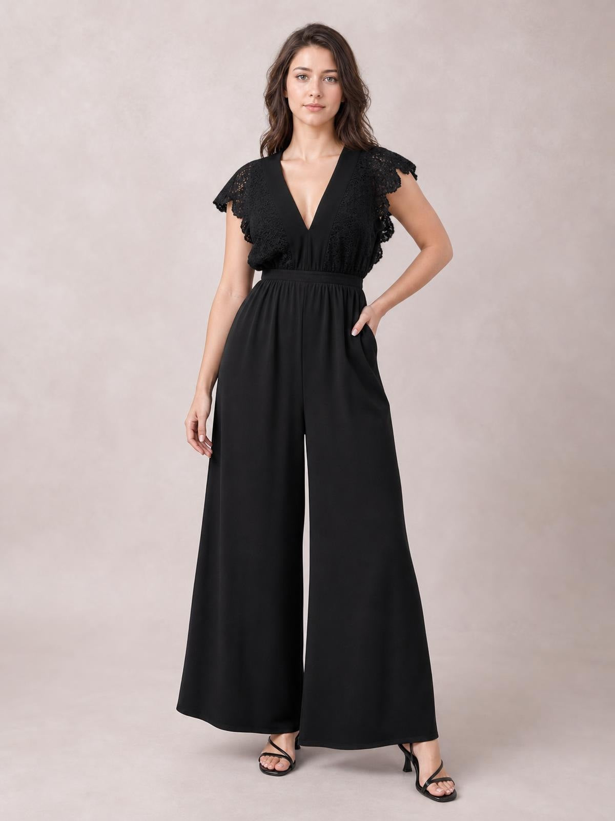 Model wearing a black lace cap sleeve jumpsuit with a deep V-neck, buttoned bodice, defined waist, and flowing wide-leg silhouette, posing with one hand in a pocket.