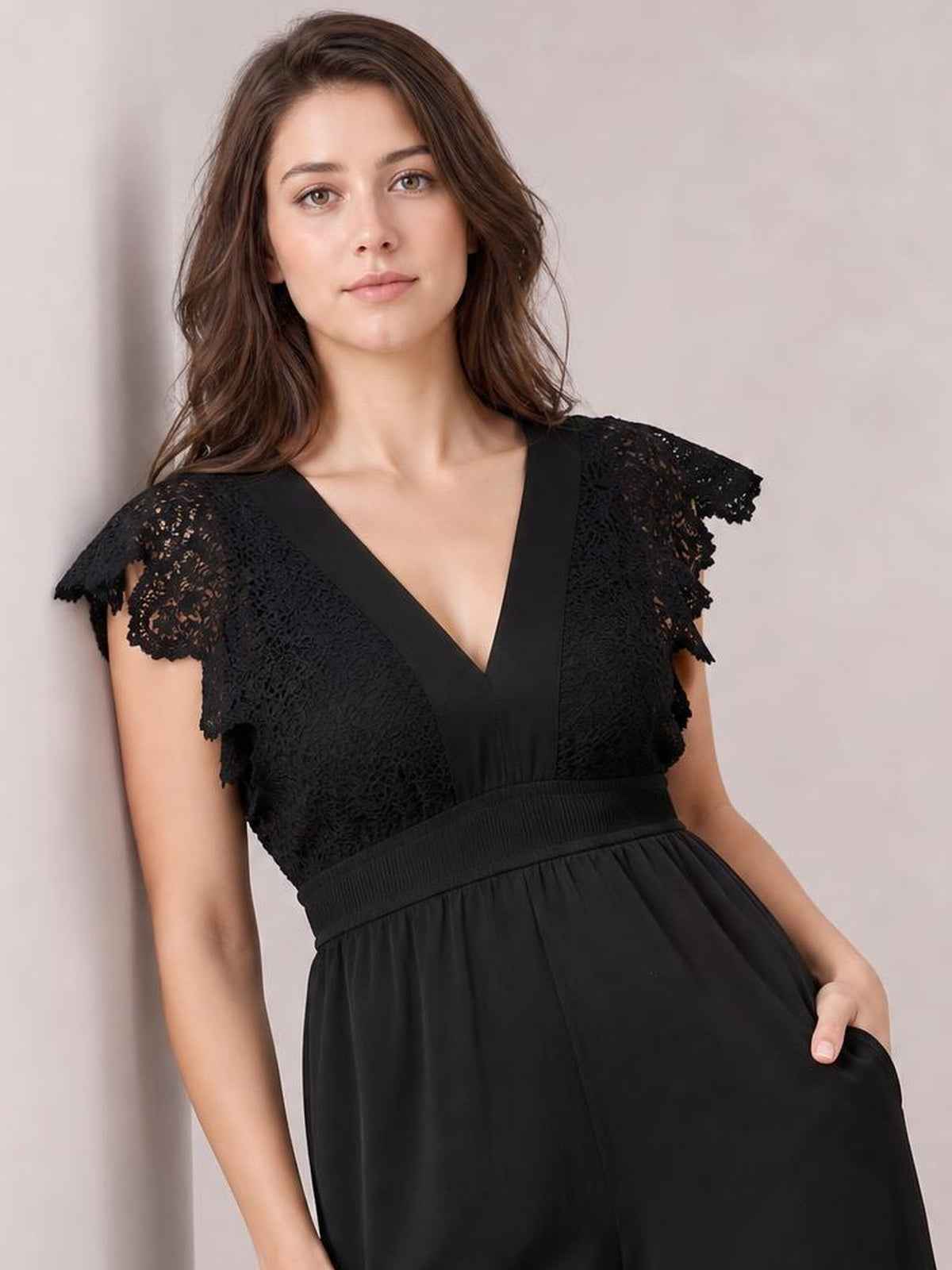 Model poses in a black lace cap sleeve jumpsuit with a V-neck, wide-leg silhouette, elasticated waist, and a hand resting in the side pocket.