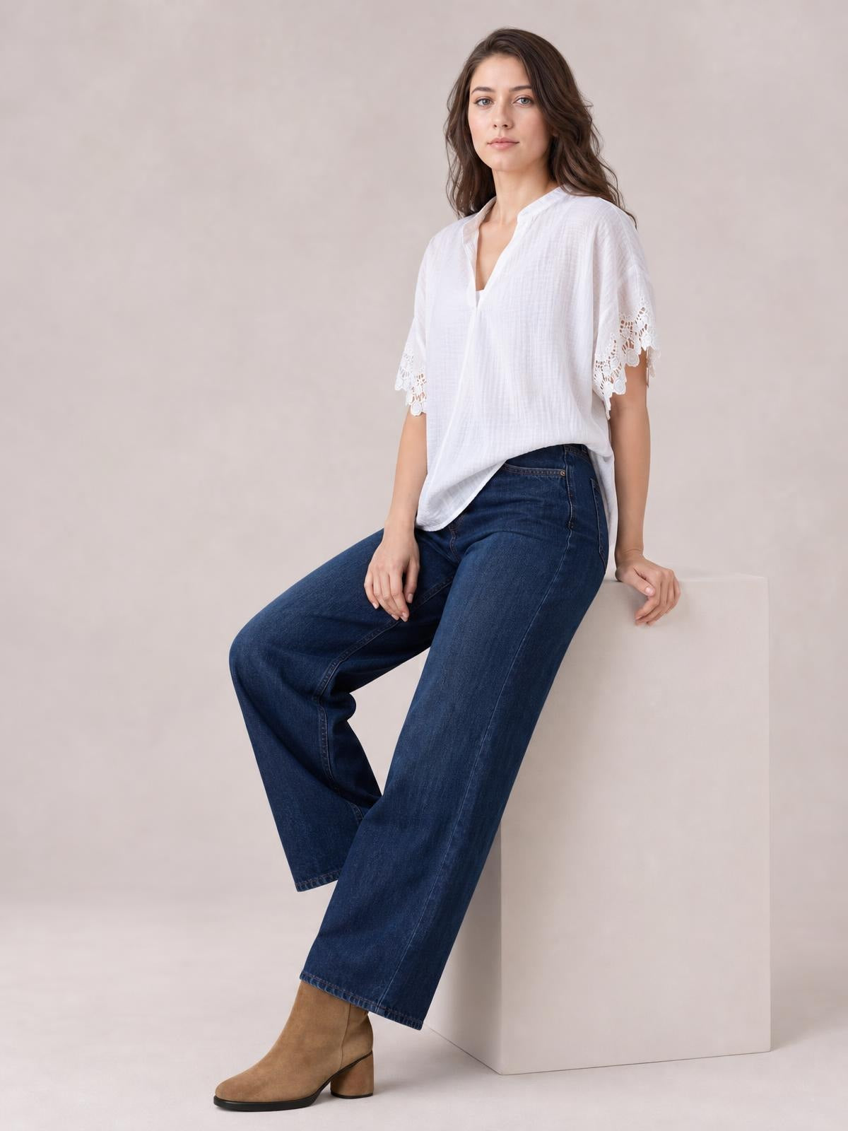 Model seated in a white collared short-sleeve blouse with lace-trimmed cuffs, a relaxed silhouette, and an open neckline, paired with wide-leg jeans.
