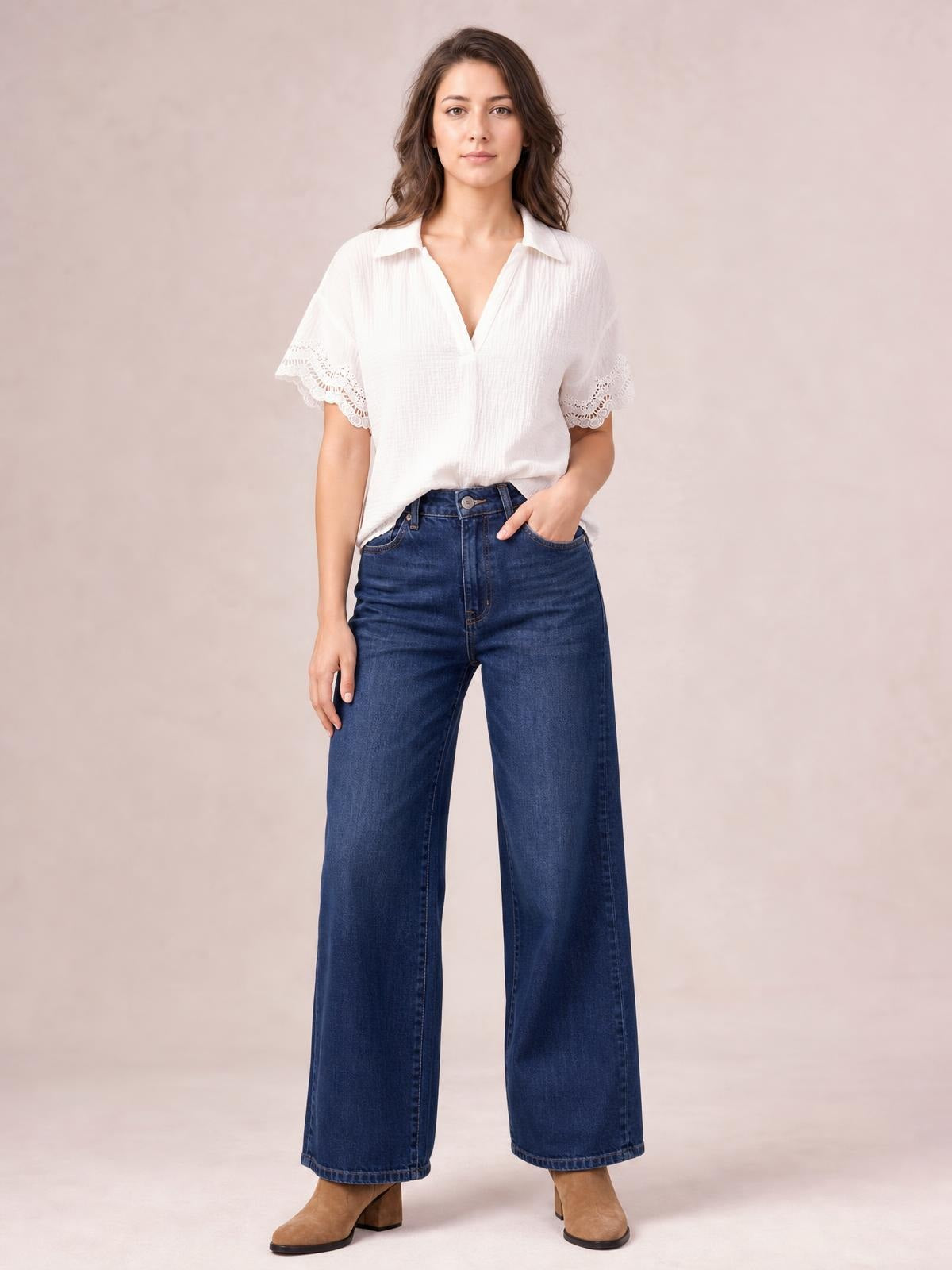 Model wearing a white collared short sleeve blouse with lace-trimmed sleeves and a relaxed silhouette, posed with one hand in her jeans pocket.
