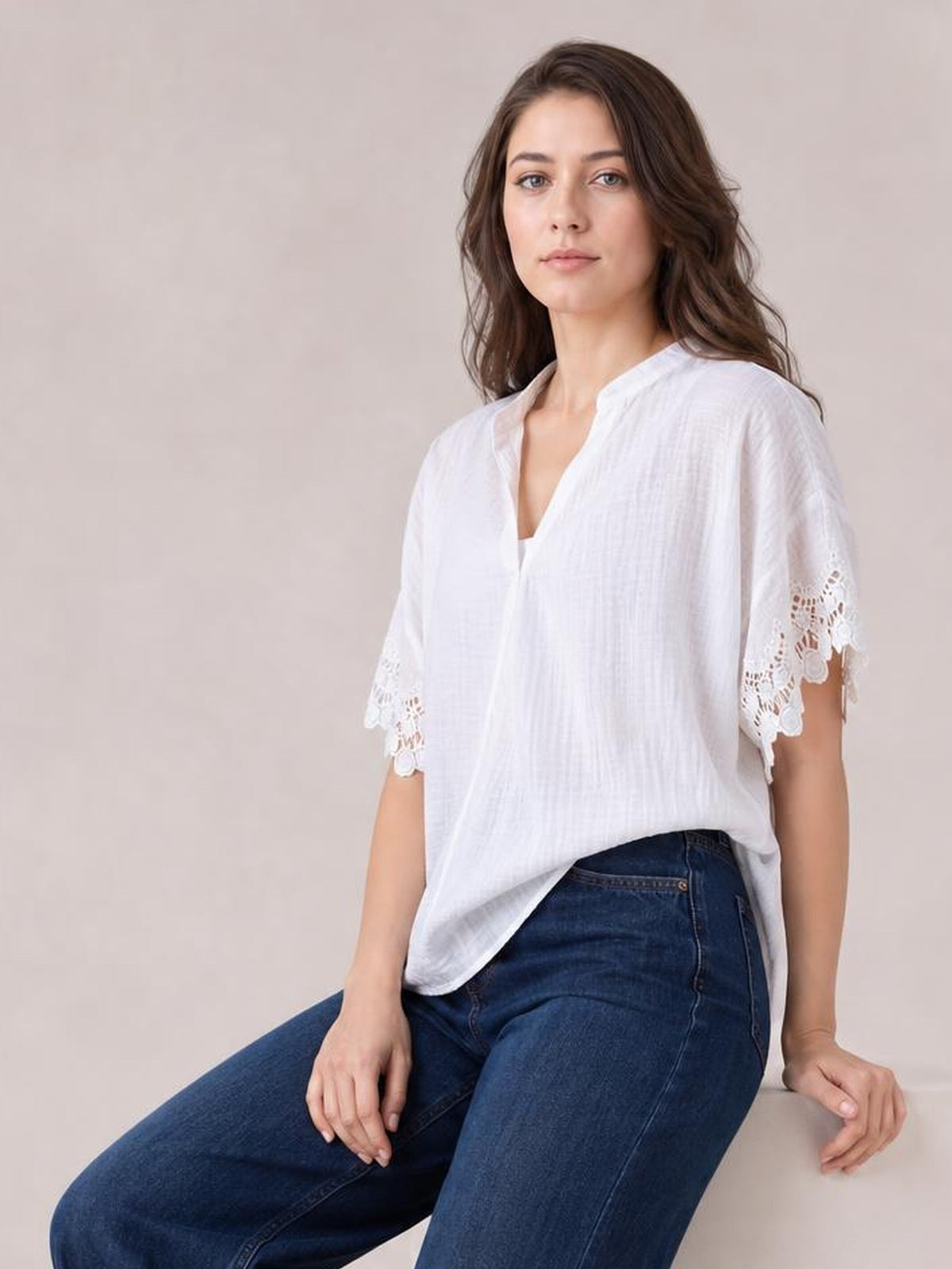 A seated woman wears a white collared blouse with a relaxed silhouette, V-neck opening, short sleeves, and scalloped lace detail at the cuffs.