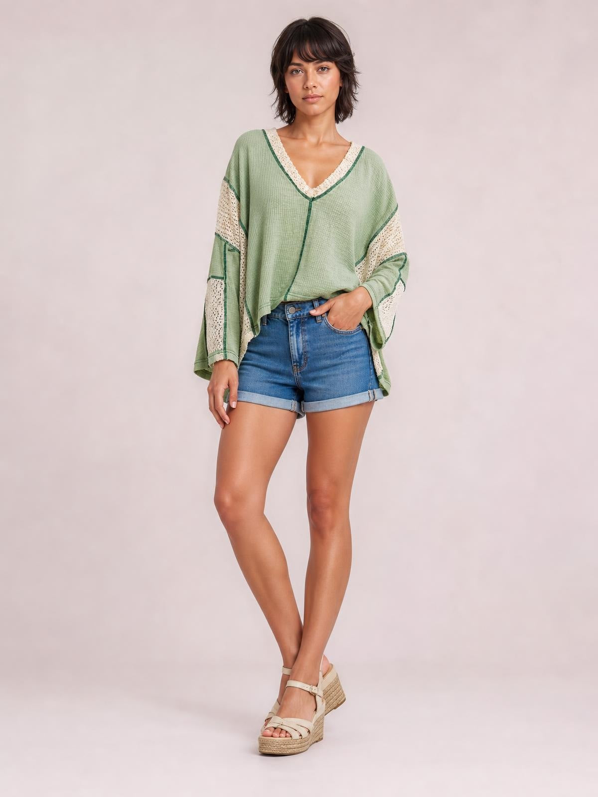 Model poses with one hand in her shorts pocket, wearing a sage green contrast V-neck long sleeve blouse with cream lace details and a relaxed silhouette.