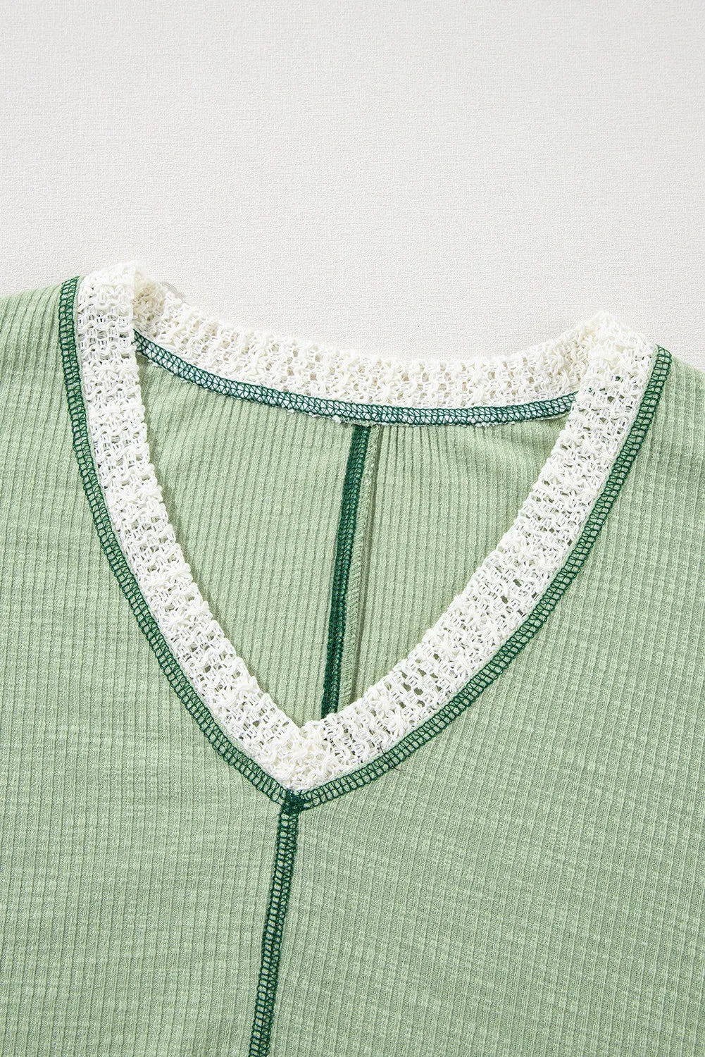 Close-up of a sage green ribbed blouse with a contrast V-neck, white lace trim, dark green edging, and a visible center seam.