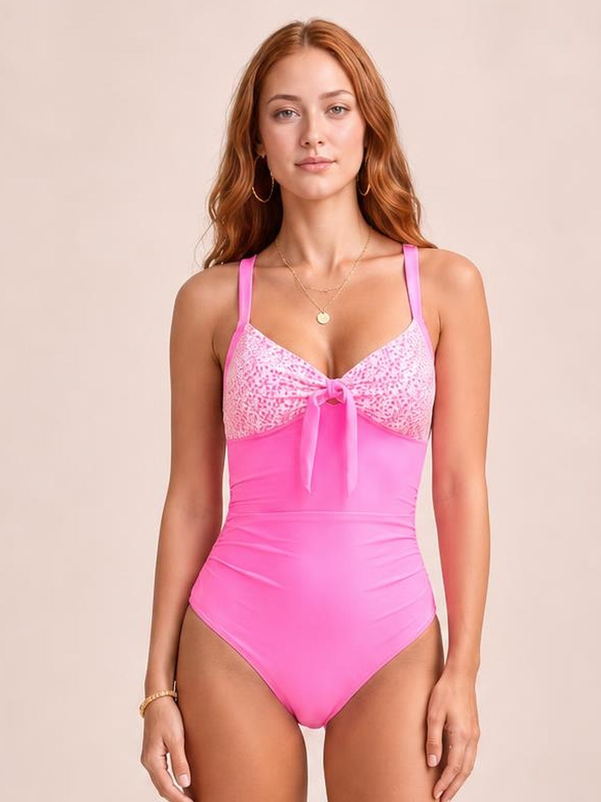 A model stands facing forward in a bright pink one-piece swimsuit with lace-detail cups, shoulder straps, a center tie, and a high-cut silhouette.