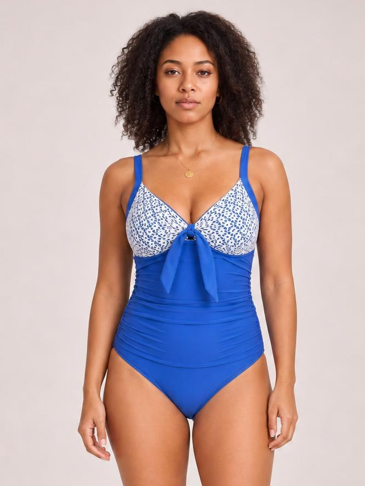 A front-facing model wears a bright blue one-piece swimsuit with lace-detail patterned cups, wide straps, a center tie, and a ruched high-waisted silhouette.