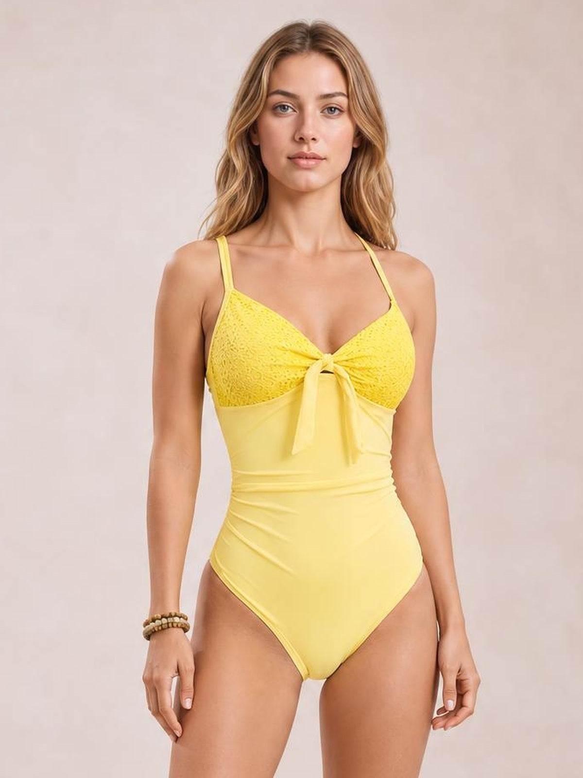 A model wears a yellow lace-detail one-piece swimsuit with thin straps, a front tie, and a fitted, high-cut silhouette.