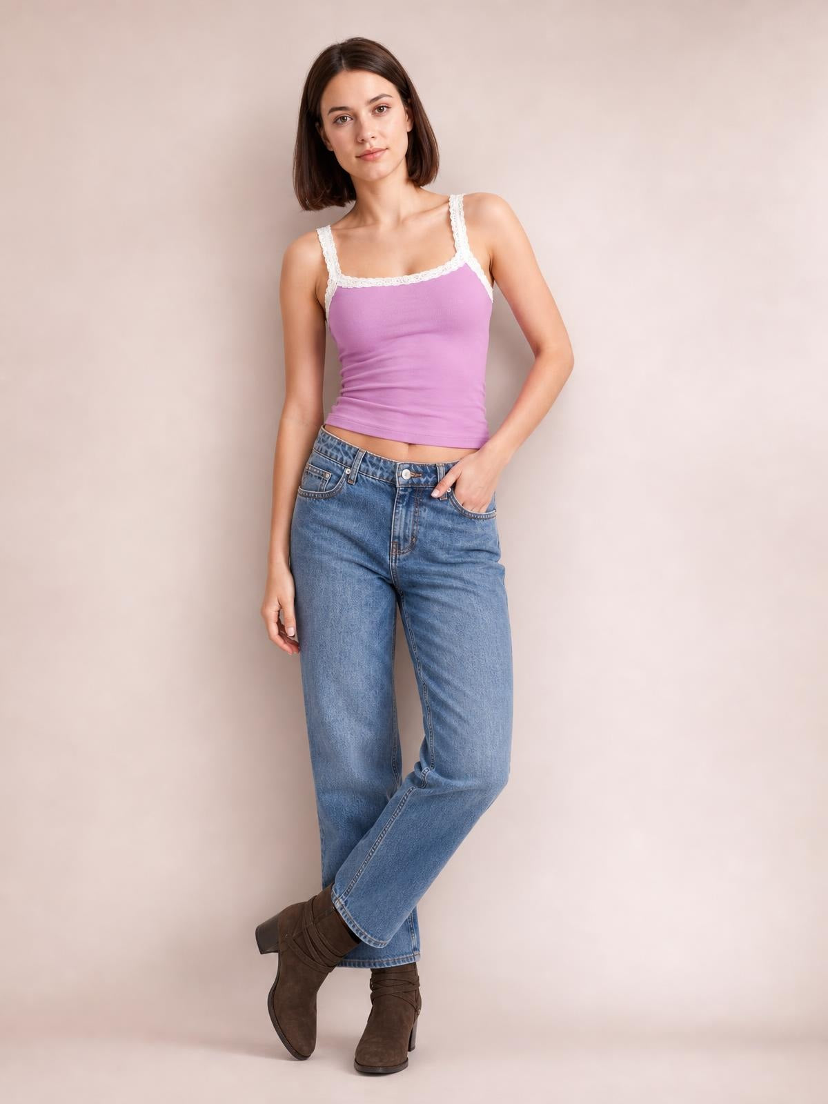 A model poses in a fitted lavender cropped cami with white lace trim along the neckline and straps, one hand in her jeans pocket and legs crossed.