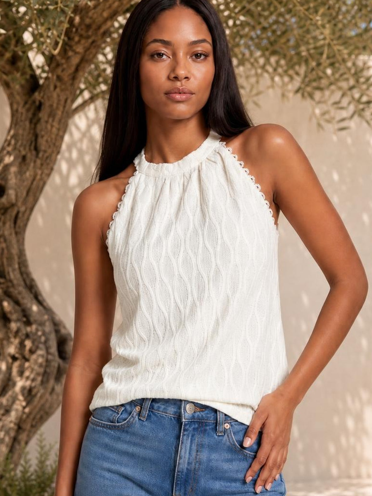 Model poses in a white Grecian neck tank with lace-trimmed edges, textured vertical detailing, a relaxed straight silhouette, and one hand in her jeans pocket.