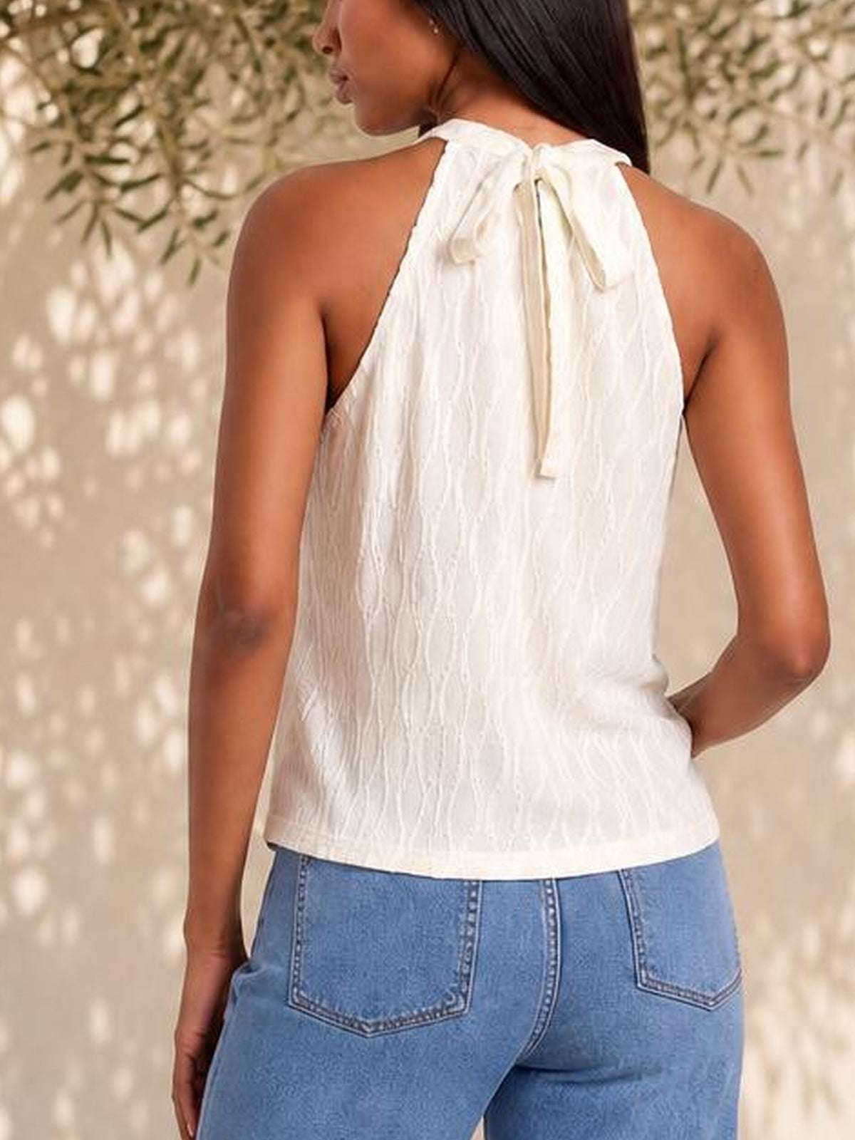 Back view of a model wearing a white lace-detail Grecian neck tank with a tied neckline and relaxed straight silhouette, posed with one hand on her hip.