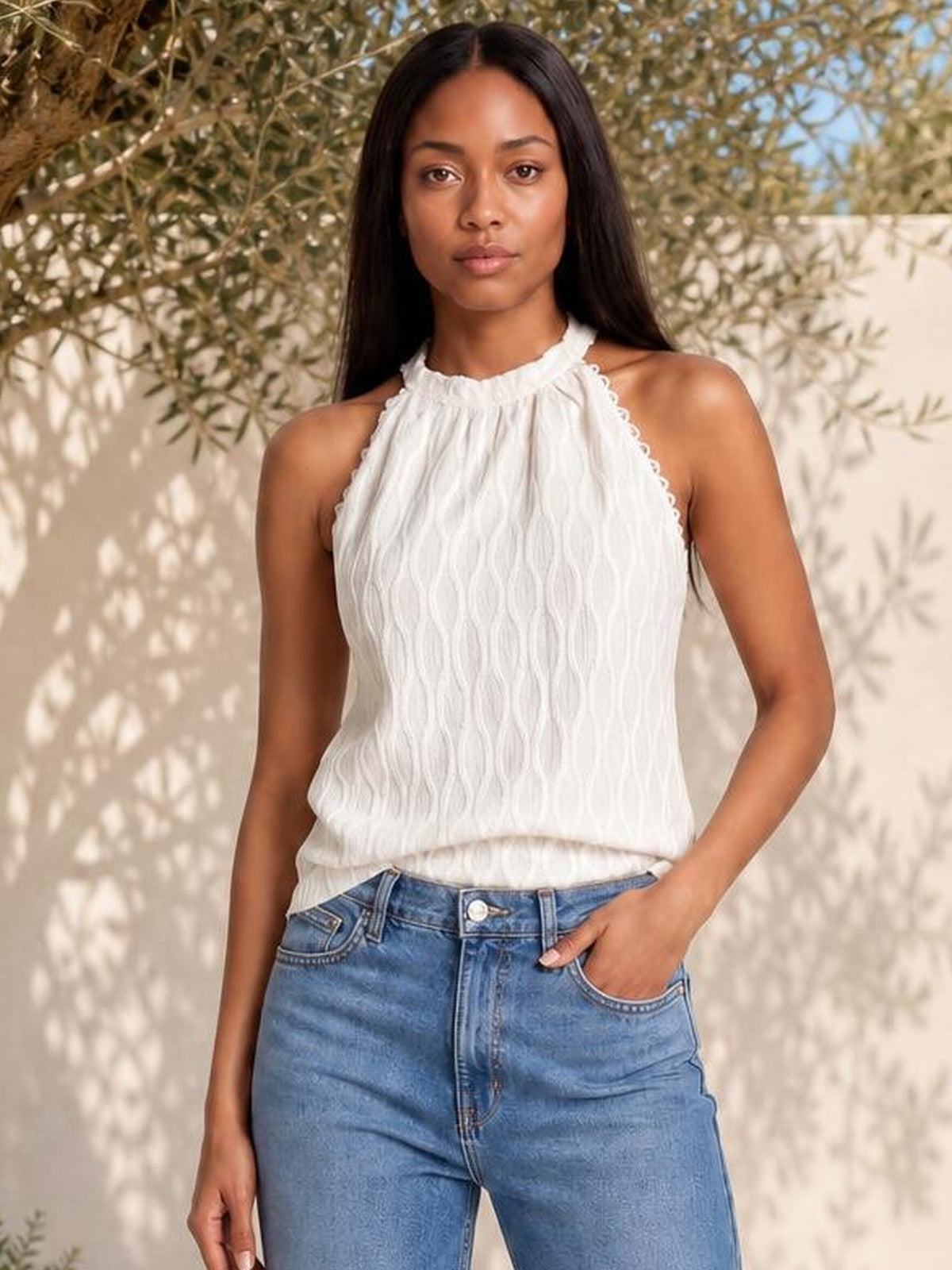 Model wears a white lace-detail Grecian neck tank with a gathered neckline, sleeveless cut, and relaxed draped silhouette, paired with blue jeans.