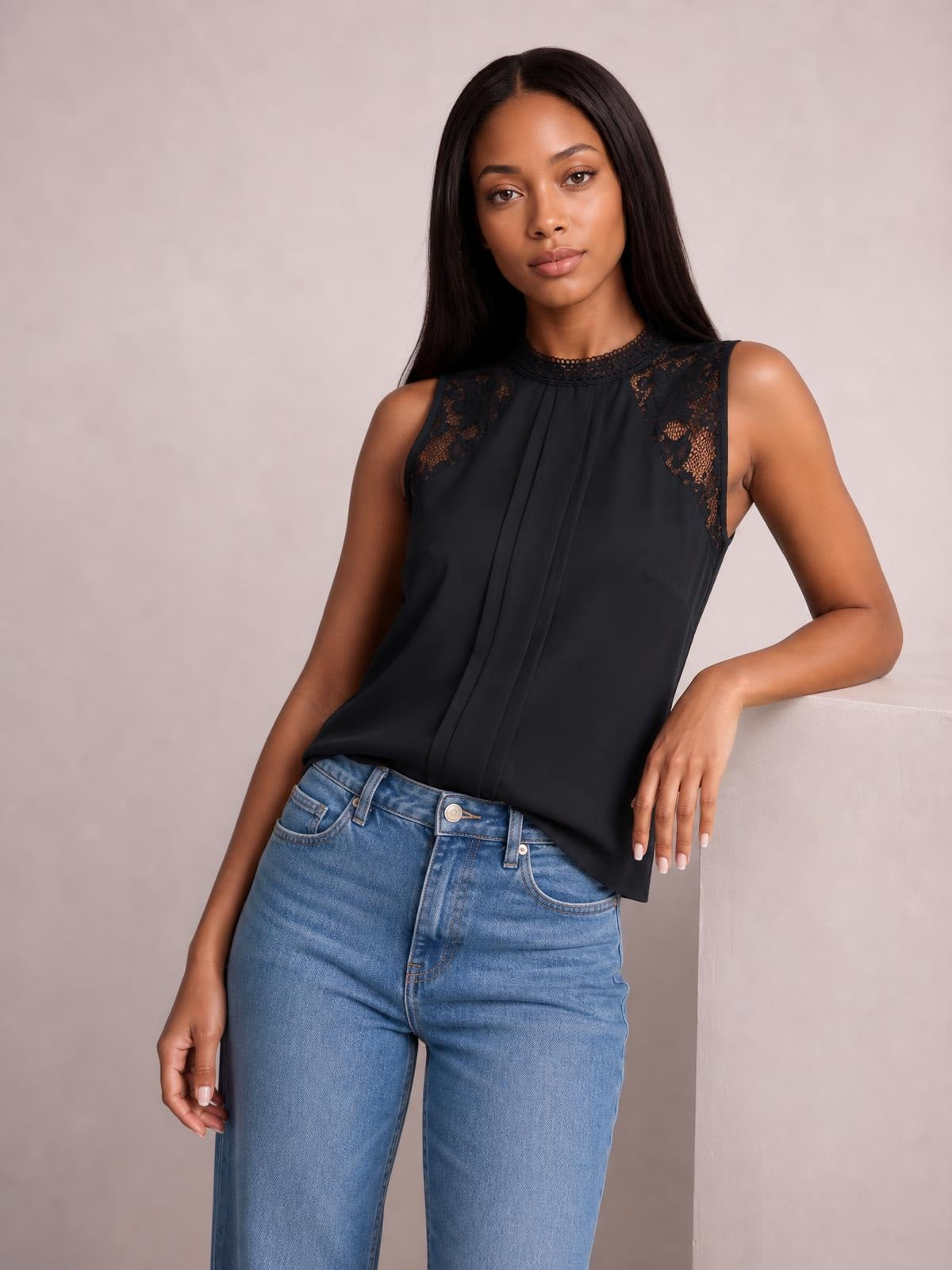 A model poses in a black sleeveless mock neck top with semi-sheer lace shoulder panels, pleated front detailing, and a relaxed straight silhouette.