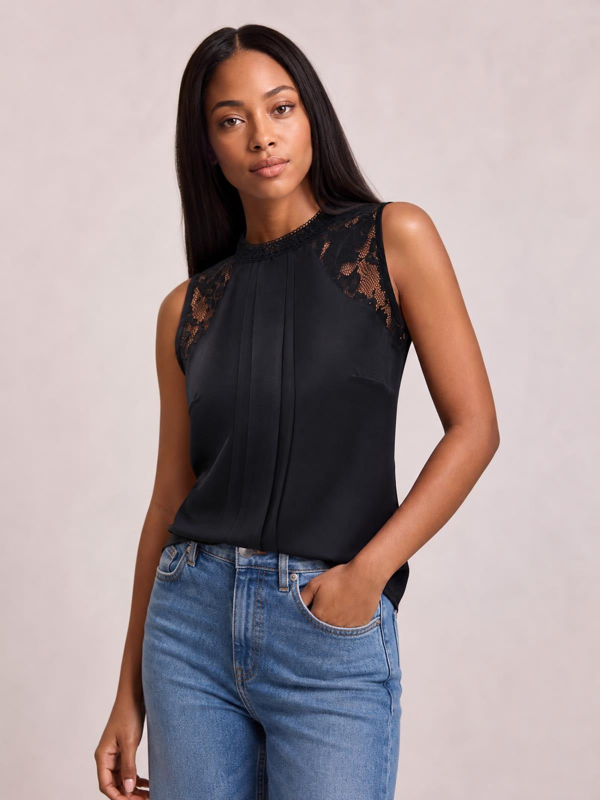 Model poses in a black semi-sheer sleeveless top with a mock neck, lace shoulder details, and a softly draped silhouette, paired with jeans.
