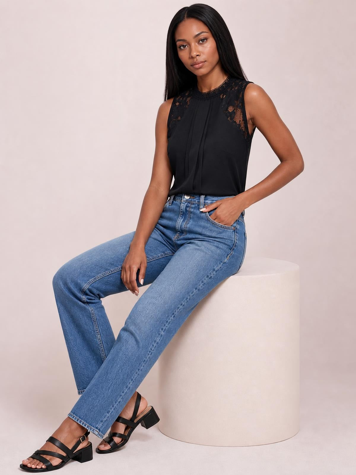 Model seated in a black semi-sheer sleeveless top with a mock neck, lace shoulder details, and a softly pleated front.