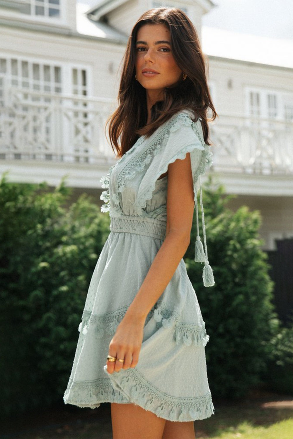 A model poses in a sage lace-detail plunge mini dress with fluttering short sleeves, a defined waist, and tiered skirt trimmed with delicate lace and ruffles.