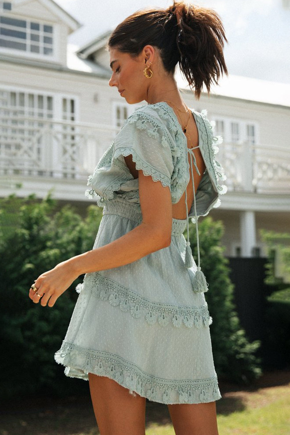 A model turns to show the sage mini dress’s open tie-back, fluttering short sleeves, lace-trimmed bodice and tiered skirt in a feminine silhouette.