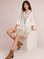 Model seated in a softly flared white printed dress with lace-trimmed neckline, gathered waist, and three-quarter bell sleeves with lace bands.