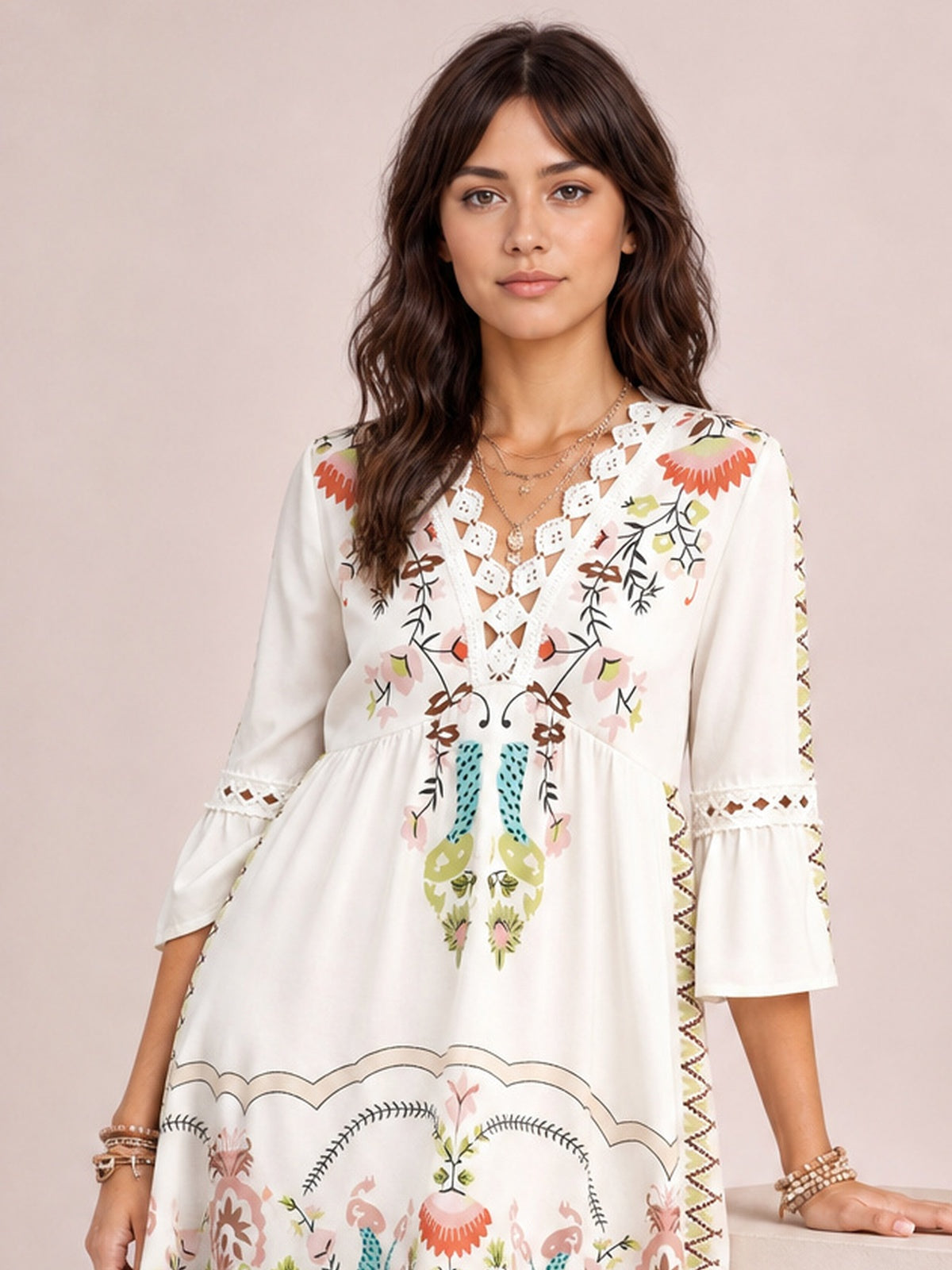 A model stands in a white lace-detail printed dress with floral motifs, a gathered waist, softly flared silhouette, and three-quarter bell sleeves with lace bands.