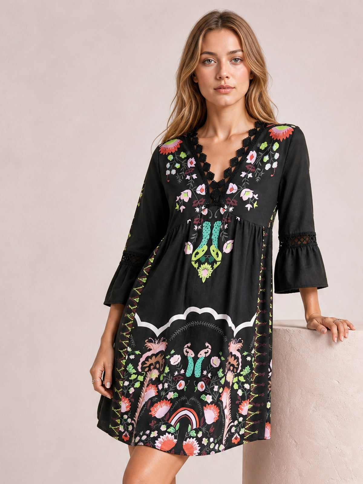 Model poses in a black printed dress with lace-trimmed V-neckline, three-quarter bell sleeves, gathered waist, and softly flared short silhouette.