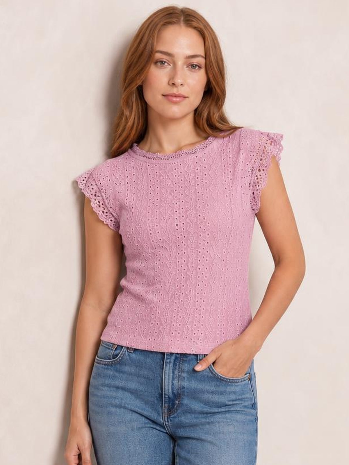 Model poses with one hand in her jeans pocket wearing a pink round-neck cap-sleeve top with eyelet lace detailing and a fitted silhouette.