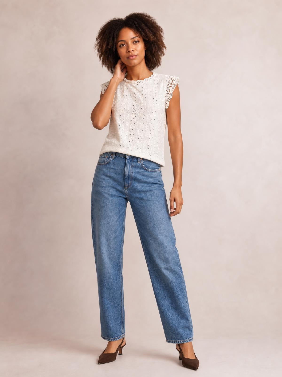 Model wearing a white lace detail round neck cap sleeve top with eyelet fabric, scalloped trim, and a relaxed straight silhouette, posing with one hand near her neck.