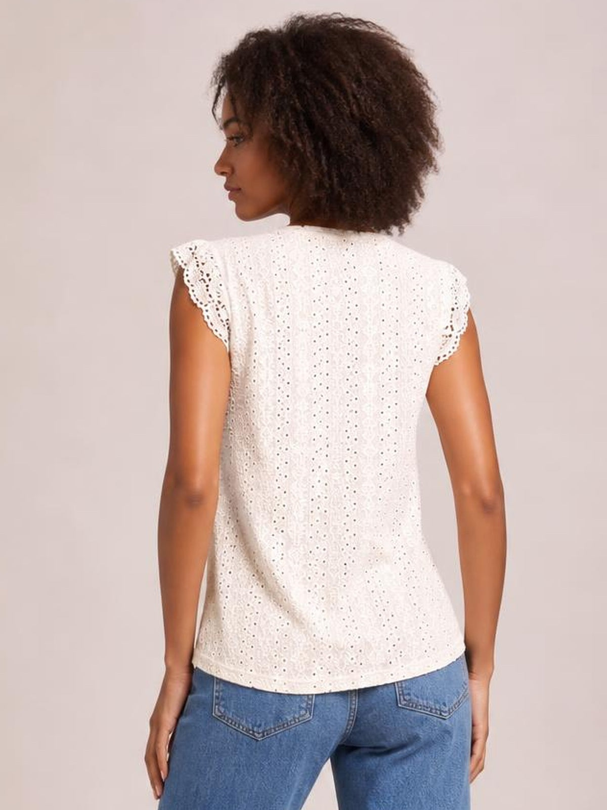 A model looks over her shoulder in a white eyelet lace top with scalloped cap sleeves and a relaxed, straight silhouette.