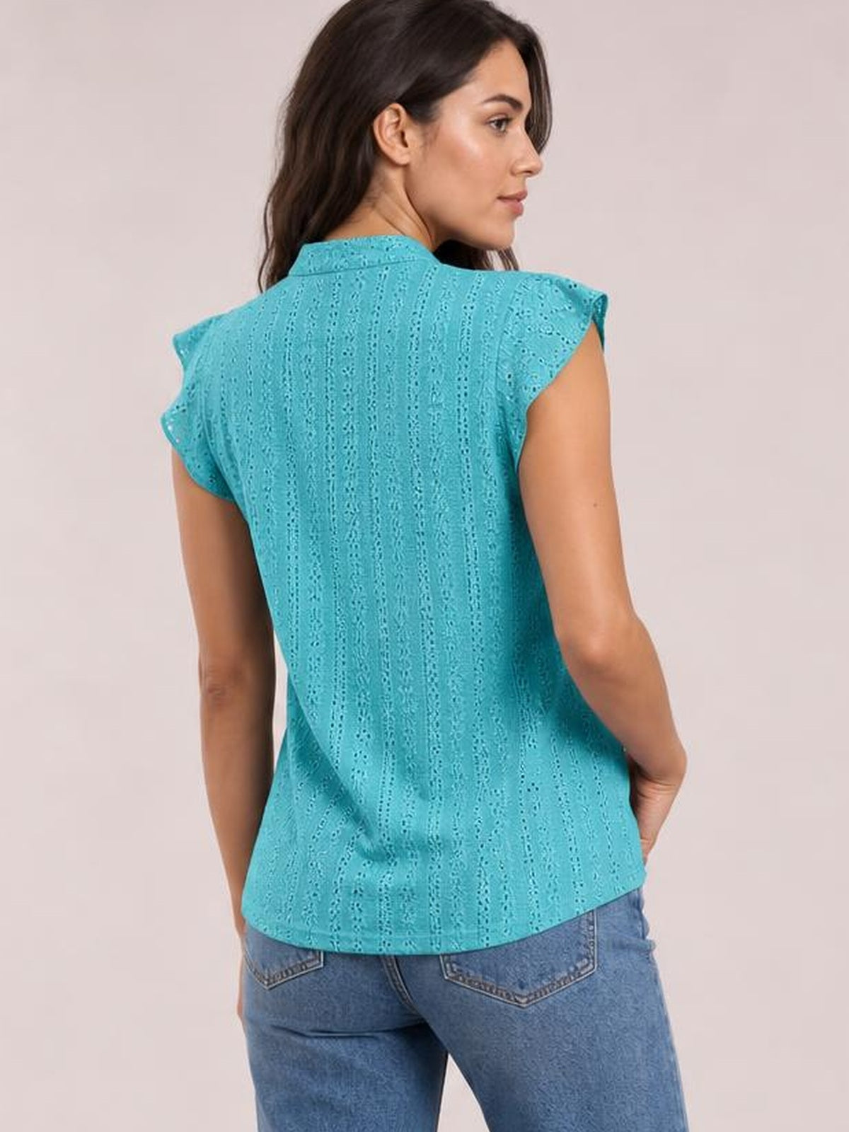 Model turns to show the back of a turquoise eyelet lace-detail top with cap sleeves, a round neckline, relaxed silhouette, and curved hem.