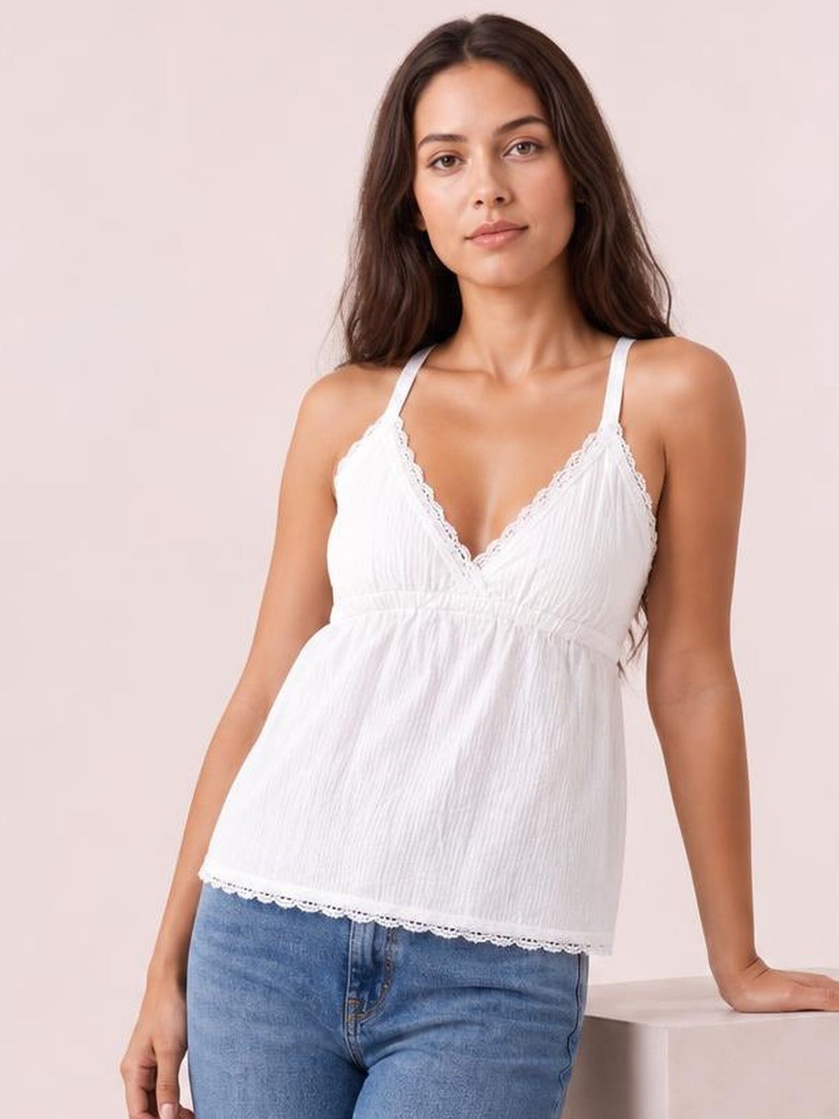 Model wears a white lace detail V-neck cami with thin straps, a gently gathered waist, and a relaxed flared silhouette over blue jeans.