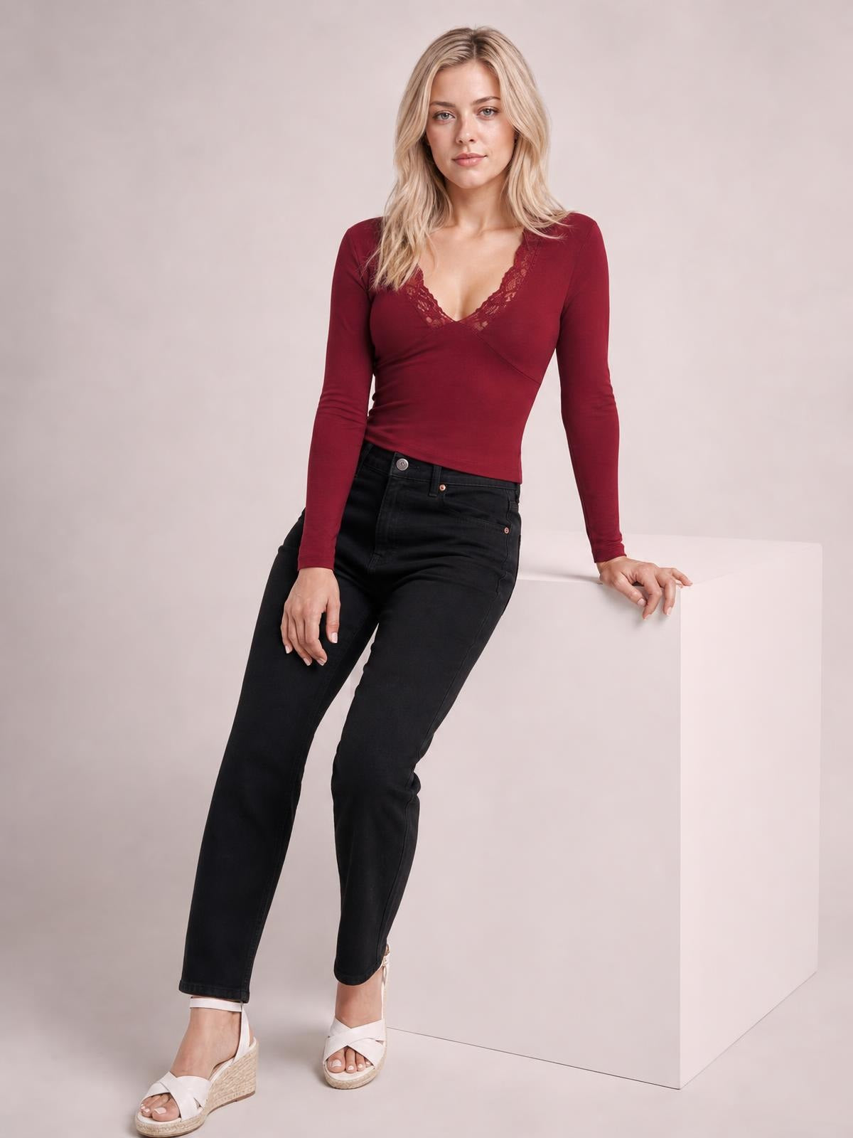 A model leans against a cube wearing a burgundy lace-detail V-neck long sleeve T-shirt with a fitted silhouette and crossover front construction.