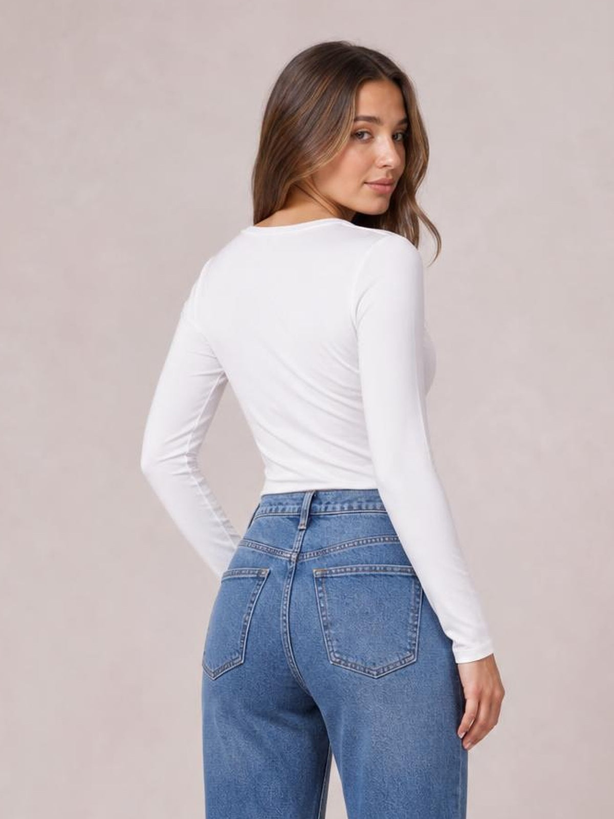 Model looks over her shoulder wearing a white opaque long-sleeve T-shirt with a fitted silhouette, paired with high-waisted blue jeans.