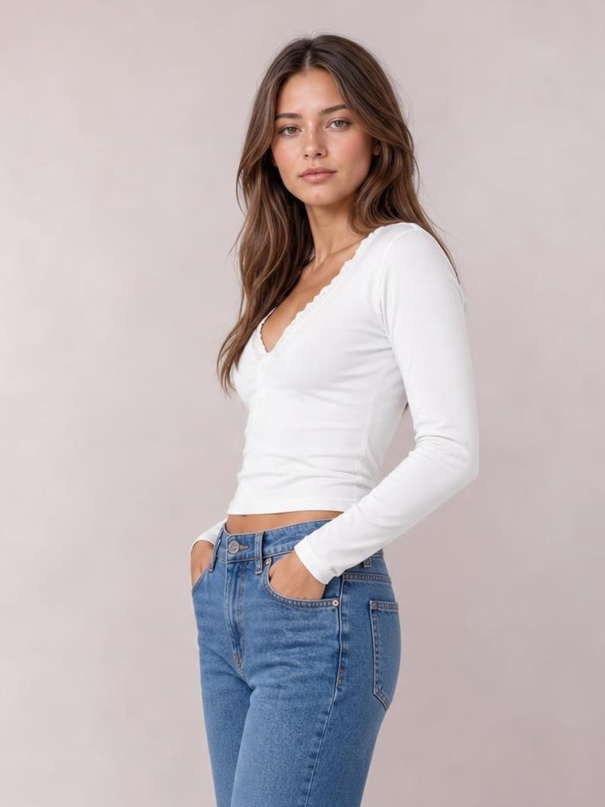 A model poses with hands in her jeans pockets, wearing a fitted white long sleeve T-shirt with a V-neckline trimmed in delicate lace.