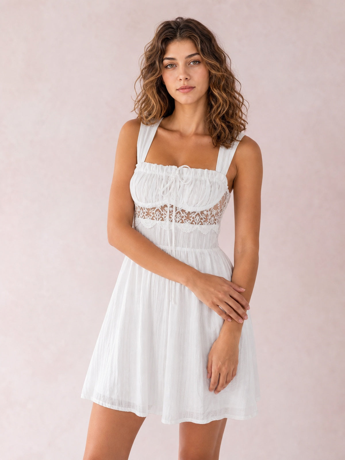Model poses in a white lace-detail mini dress with wide straps, a ruched smocked bodice, waist lace band, tie accent, and gathered A-line skirt.