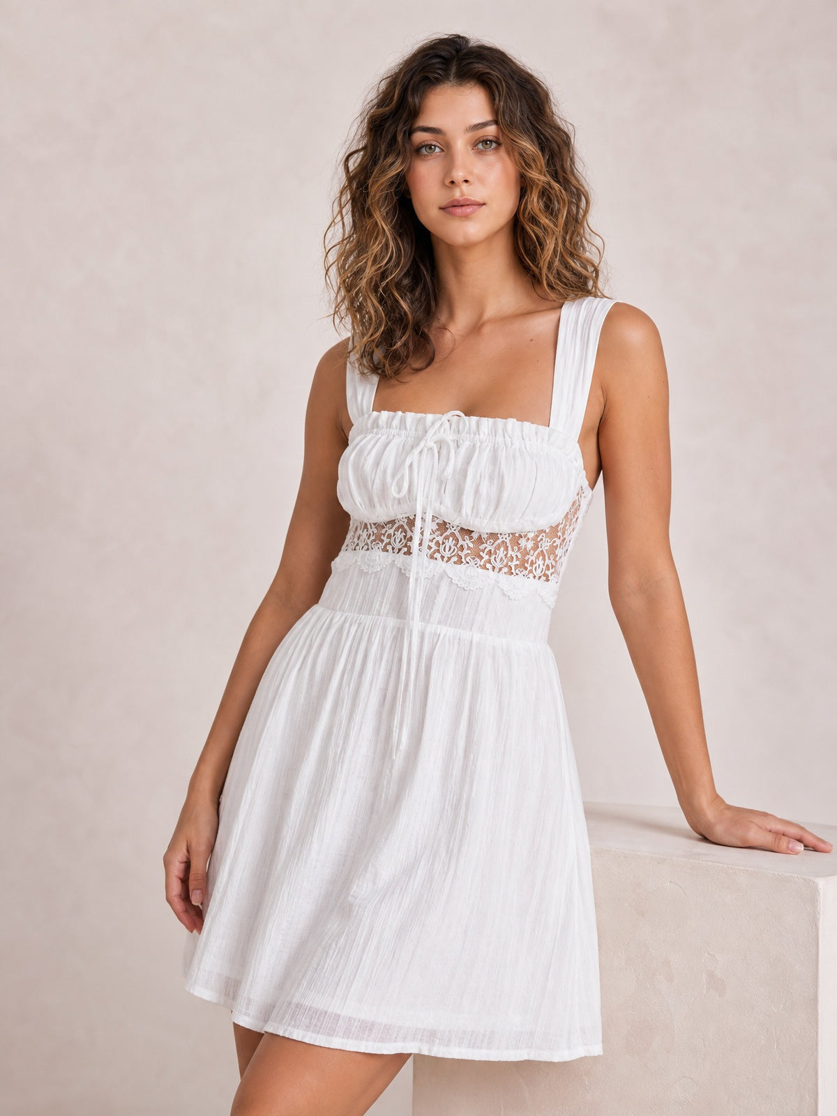 Model poses in a white lace detail mini dress with wide straps, ruched smocked bodice, tie front, defined waist, and gathered A-line skirt.