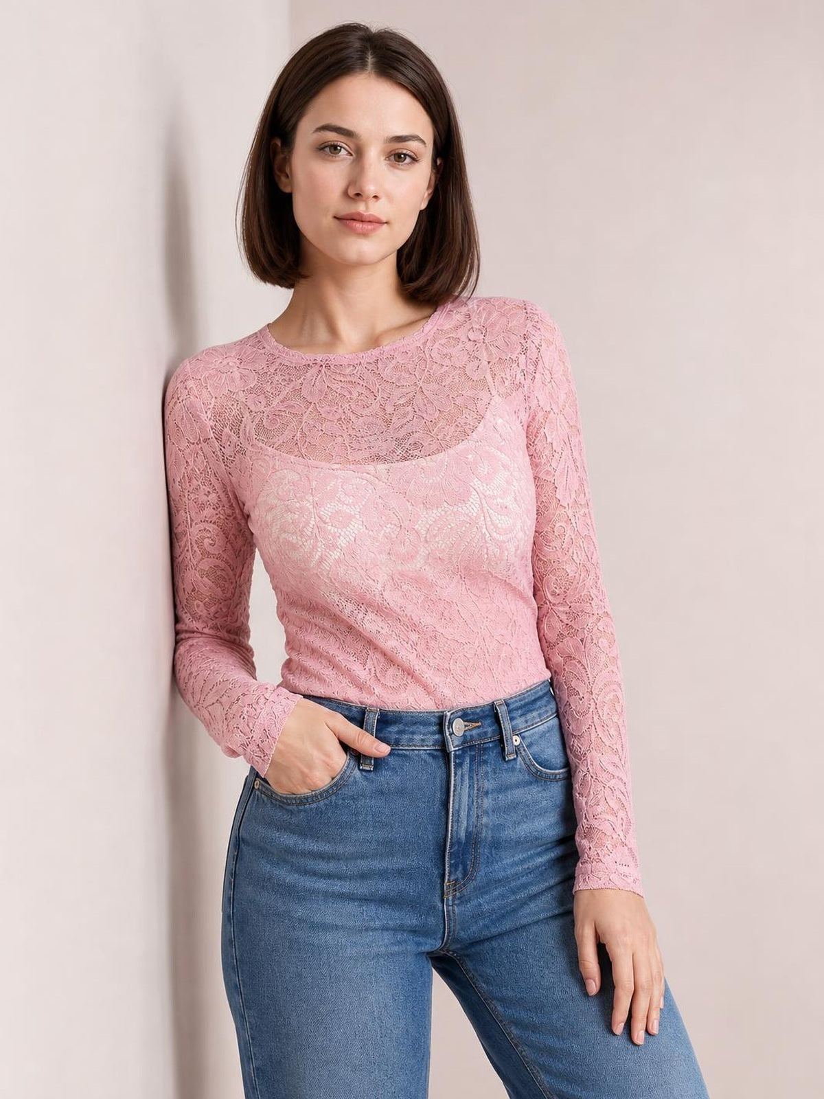 Model wearing a pink sheer lace flower top with a round neck, long sleeves, and a fitted silhouette, posing with one hand in her jeans pocket.