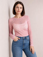 Model wearing a pink sheer lace flower top with a round neck, long sleeves, and a fitted silhouette, posing with one hand in her jeans pocket.