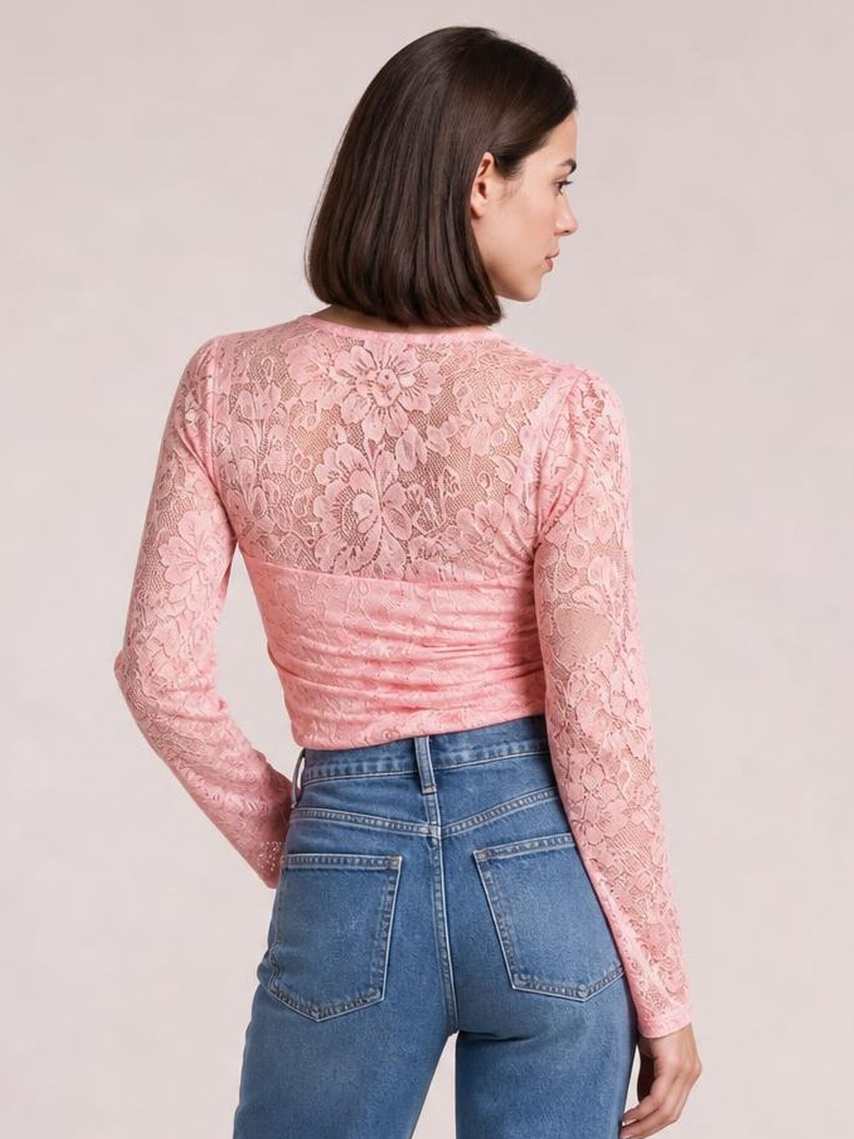 A model poses in a pink sheer floral lace top with a round neckline, long sleeves, fitted silhouette, and slightly cropped hem.