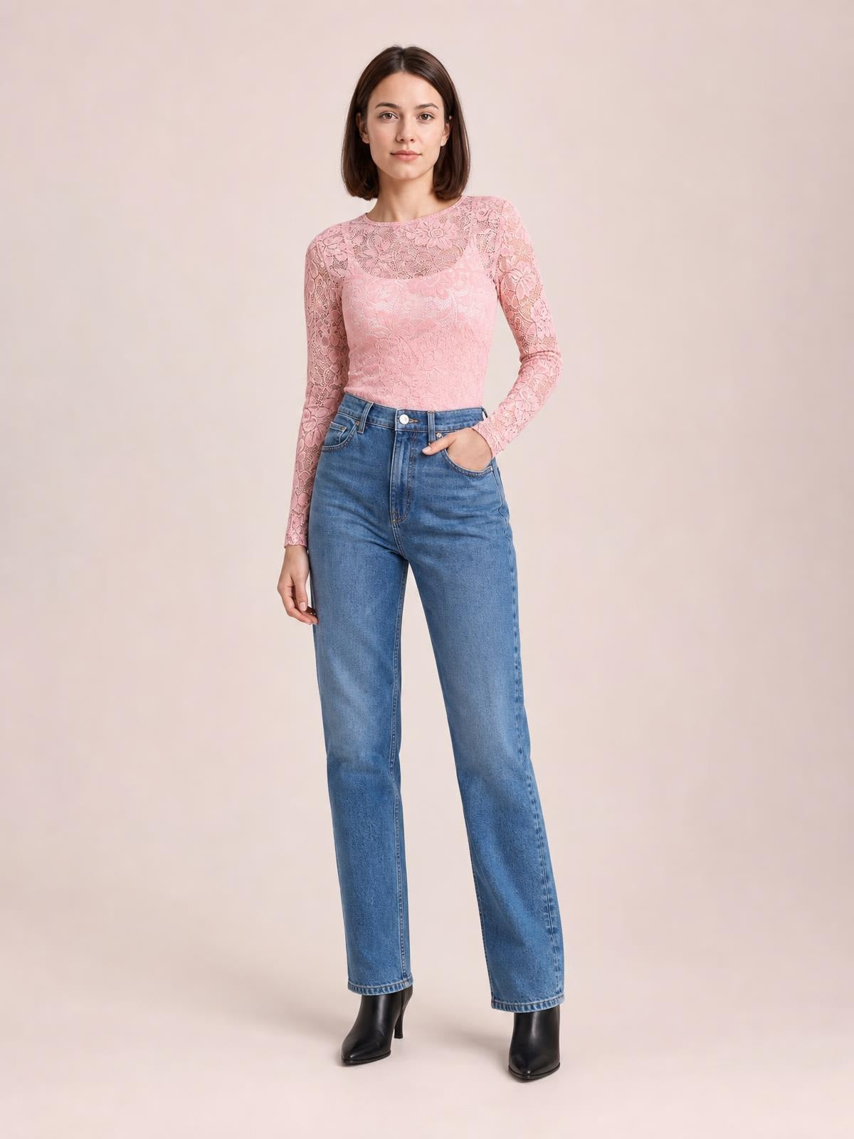 Model poses in a pink sheer floral lace top with a round neck, long sleeves, fitted silhouette, and slight stretch, resting one hand in her jeans pocket.