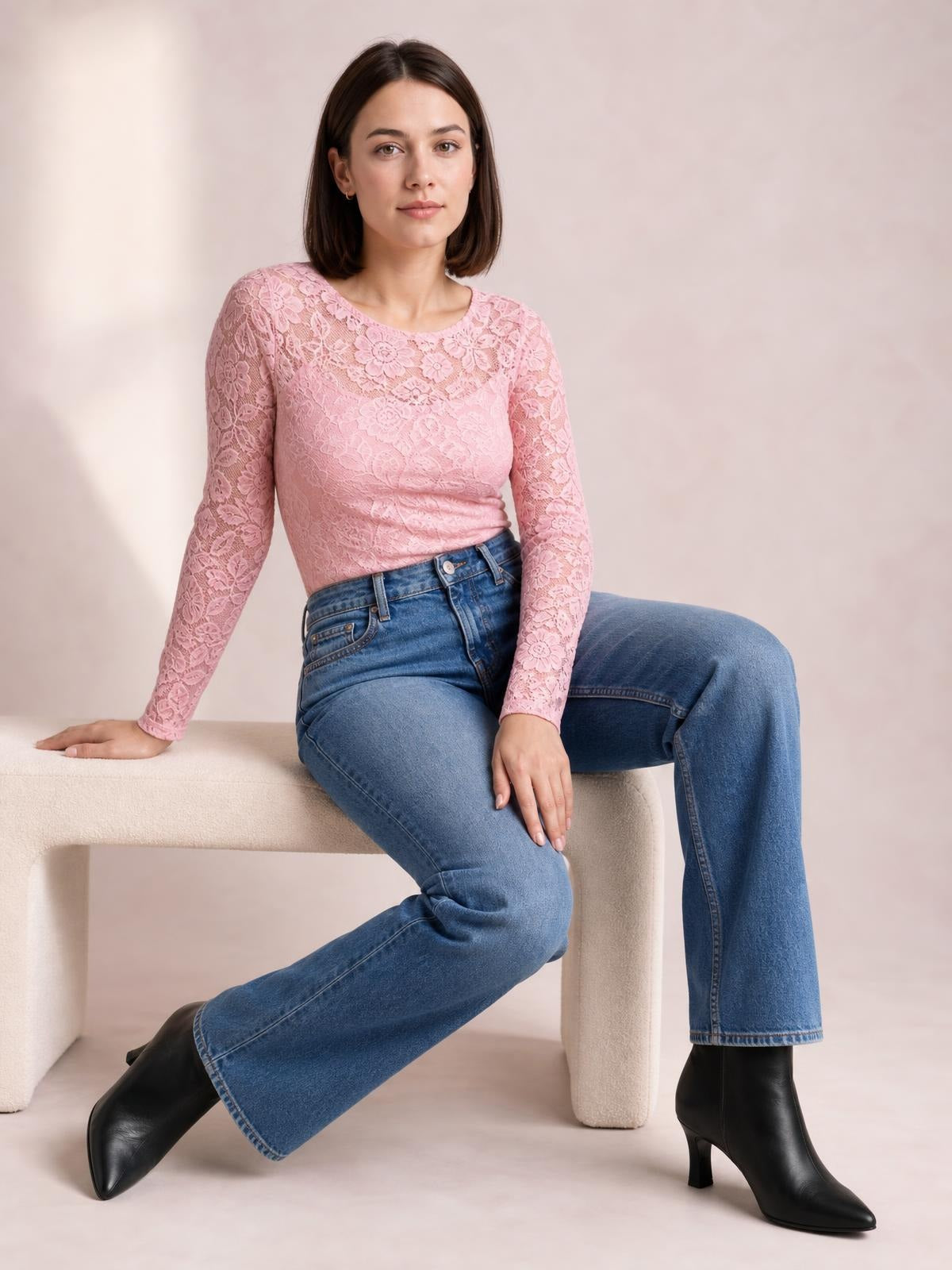 A seated model wears a pink sheer floral lace top with a round neck, long sleeves, and a fitted silhouette with slight stretch.