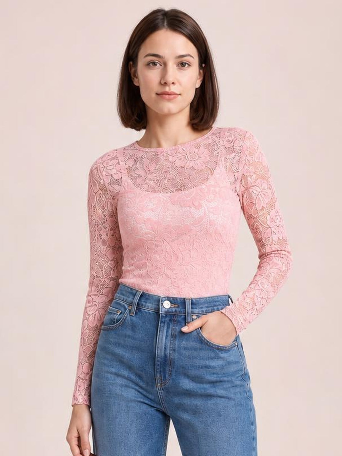 Model wearing a pink sheer lace flower top with a round neck, long sleeves, fitted silhouette, and floral lace construction, posed with one hand in her jeans pocket.