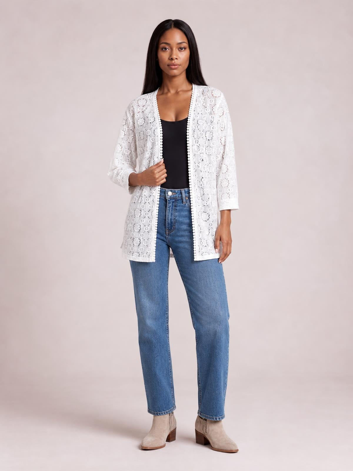 Model stands in blue jeans wearing a white lace open-front cover-up with three-quarter sleeves, a relaxed silhouette, and scalloped trim, holding one side.